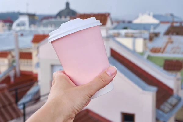 Pink paper cup with coffee in a woman's hand. It's time to drink coffee in the city. Disposable paper cup close-up on the background of the city. Empty space for text, layout. Coffee to go concept
