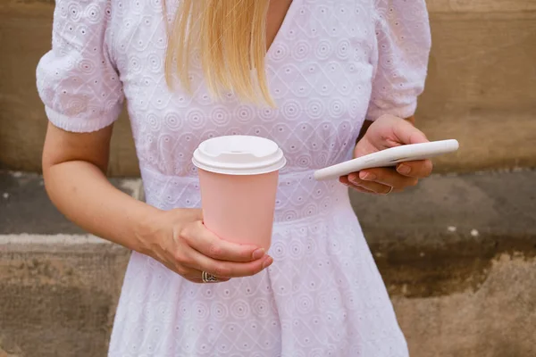 Girl drinks coffee from a pink paper cup and uses a smartphone. A woman is holding a smartphone, sending a text message, or using an app on her mobile phone. Drink cappuccino or latte in the street