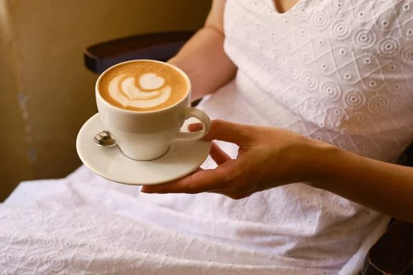 A woman with a cup of aromatic coffee in her hands sits in a cafe or restaurant. The girl drinks coffee after breakfast. Delicious hot latte or cappuccino in a white cup. Quiet freelance lifestyle