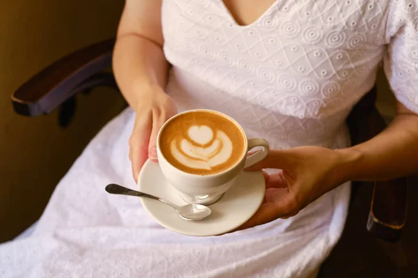 A woman with a cup of aromatic coffee in her hands sits in a cafe or restaurant. The girl drinks coffee after breakfast. Delicious hot latte or cappuccino in a white cup. Quiet freelance lifestyle