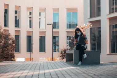 Maskeli genç kahverengi saçlı öğrenci teneffüste üniversite sınavını geçmek için ders çalışıyor.