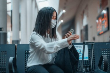 Otobüs durağında maskeli genç bir kız yanında seyahat çantasıyla bekliyor.