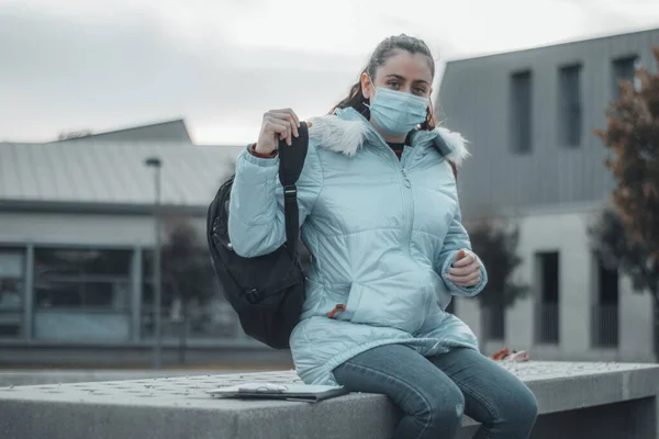 Joven estudiante quitándose la mochila con una máscara mientras está sentada en el centro universitario para descansar en el recreo y tomar notas — Foto de Stock