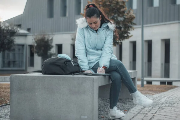 在大学校园里，一个年轻的女学生坐在长椅上学习笔记和书本 免版税图库图片