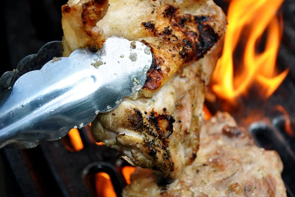 Szczypce trzymając kawałek kurczaka z ogniem grilowania w baackground — Zdjęcie stockowe
