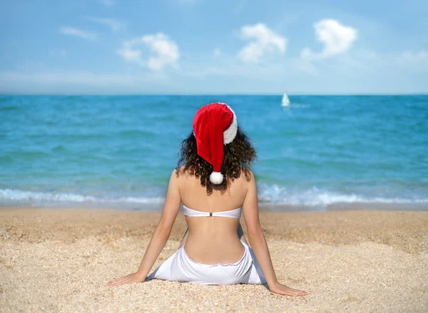 Молода дівчина носити капелюх Санта Клауса, сидячи на пляжі і дивитися на море — стокове фото