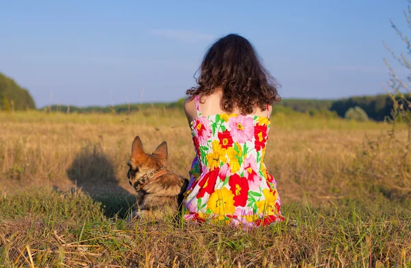 젊은 여 자가 석양 초원에 카메라를 다시 그녀의 강아지와 함께 앉아. — 스톡 사진