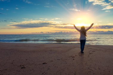 Güneş doğarken deniz manzarası ve güzel bir gökyüzü. Elleri havada genç ve mutlu bir kadın sahilde duruyor ve güneşin doğuşunu seyrediyor.