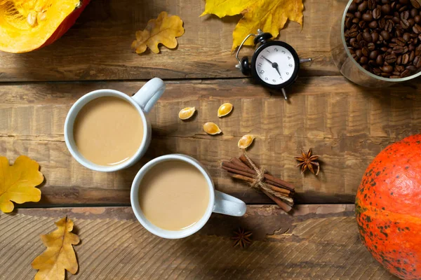 Two cups of pumpkin coffee with spices, orange pumpkins, dry oak leaves and an alarm clock on a wooden background. Autumn mood. Time to drink hot beverage. Top view.