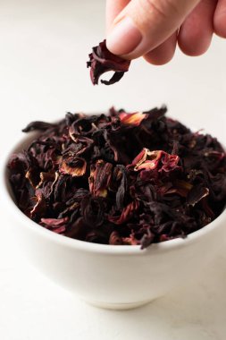 Beyaz bir kâsede kurumuş amber yaprağı uzanır. Beyaz arka planda bir kadının eli bir yaprak kaldırır. Kurutulmuş rosella ya da Sudan gülü çiçeği. Arap ülkelerinde çok popülerdir. Dikey yönelim.