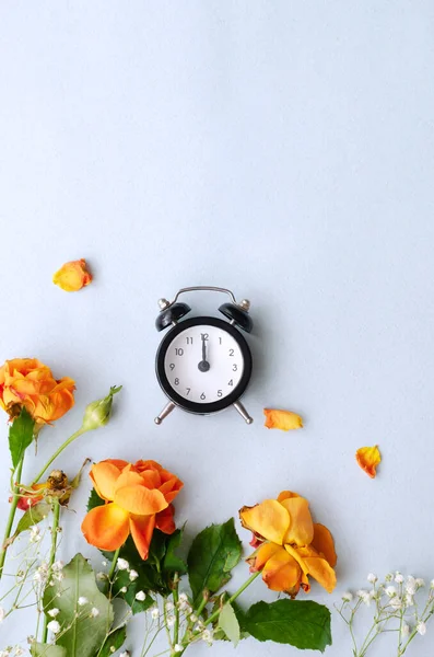 Düşen yapraklı turuncu güller ve mavi arka planda siyah bir saat. Zaman geçirme kavramı. Eski çiçekler. Şık değil. Dikey oryantasyon. Üst görünüm.