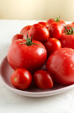 Farklı boyutlarda kırmızı domatesler. Beyaz zemin üzerinde pembe büyük bir tabakta su damlaları var. Vejetaryen yemeği kavramı. Dikey yönelim.