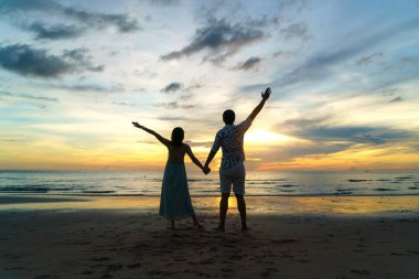 Romantik Asyalı çift ayakta ve el ele tutuşup tatil boyunca sahilde gün batımının tadını çıkarıyorlar..