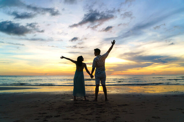 Романтическая азиатская пара, стоящая и держащаяся за руку, наслаждаясь прекрасным закатом на пляже во время отпуска во время путешествий.