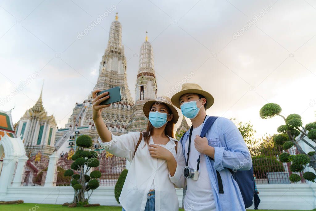 Asian couple happy tourists to travel wearing mask to protect from Covid-19 on they holidays and selfie by camera in Wat arun in Bangkok, Thailand