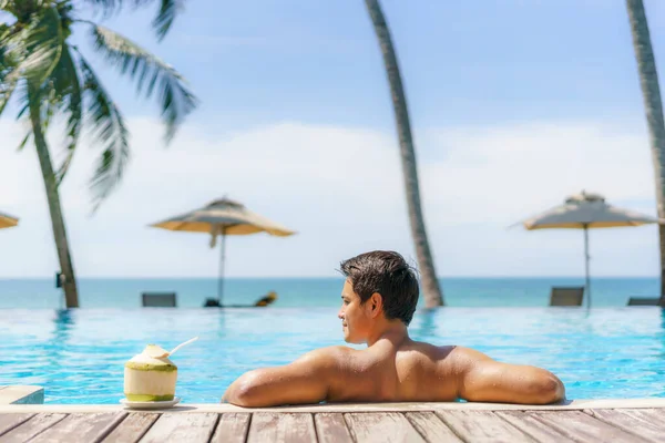 夏の旅行休暇のコンセプト ココナッツと旅行者のアジア人男性は 海のビーチとヤシの木の背景を持つ高級インフィニティプールホテルリゾートでリラックス — ストック写真