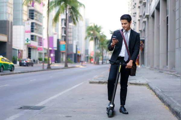 アジアのビジネスマンは 電動スクーターに乗っていると朝に動作するように都市の道路ルートを確認するためにアプリケーションで地図を開くために彼の携帯電話を使用しています — ストック写真