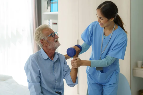 Senior man physiotherapy exercises with Asian woman caregiver or nurse physiotherapist in uniform helping aged male patient at home