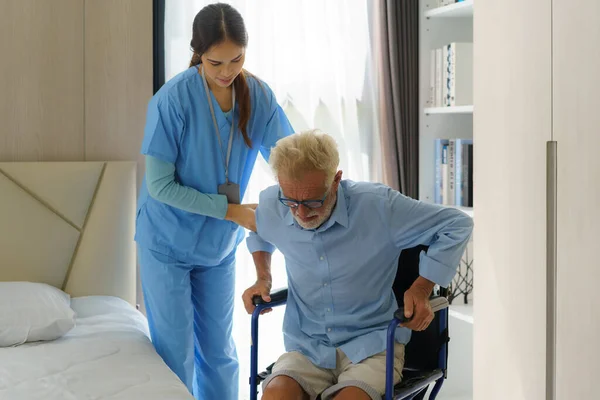 Asian smiling nurse helping senior man get out of bed nursing care support patient while getting out of bed and moving to wheelchair at home helping handicapped elderly stand up