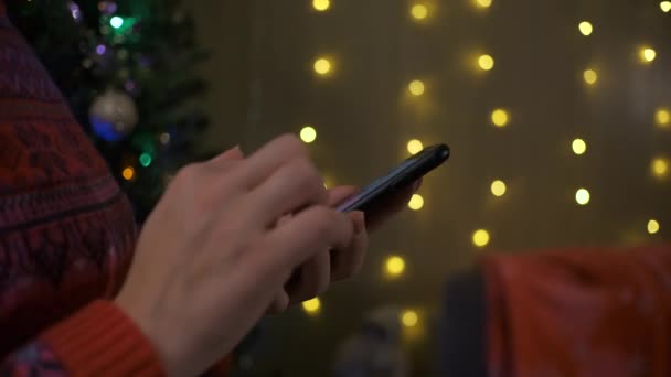 Manos con teléfono inteligente en luces borrosas del árbol de Navidad y la chimenea. Orden de regalo — Vídeo de stock