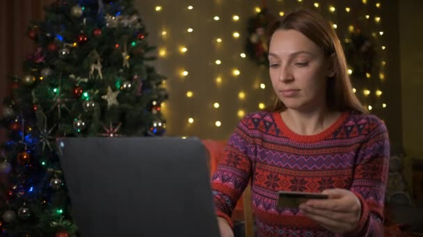 Молодая женщина лежит под елкой и заказывает подарки в интернет-магазине на ноутбуке — стоковое видео