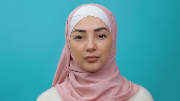 A close-up view of a beautiful serious arabian Young Muslim woman in hijab is thinking about something standing isolated over a blue background in studio — Stock Video