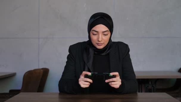 Jovem muçulmana no hijab jogando jogo móvel no ciberespaço no café. Conceito de entretenimento, compartilhamento social, vida online e Metaverse. Cultura islâmica tradicional — Vídeo de Stock