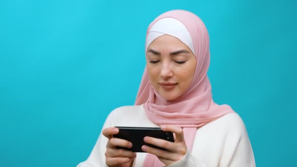 Aplicación de entretenimiento móvil. Mujer musulmana joven en hijab jugando con impaciencia videojuego en el teléfono móvil, disfrutando de la jugabilidad en tirador o carreras. estudio aislado interior plano sobre fondo azul — Vídeos de Stock