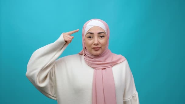 Displeased annoyed Young Muslim woman in hijab showing stupid gesture, accusing crazy cuckoo mind, blaming insane idea, reckless plan, senseless dumb talk. indoor isolated on blue background — Vídeo de stock