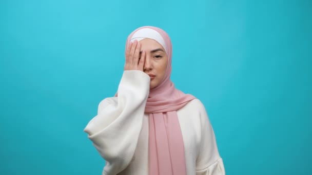 Twarz, smutne emocje. Zdenerwowana młoda muzułmanka w hidżabie uderza się w twarz i wyraża żal, obwinia się za kłopoty, czuje się bezradna. Studio nakręcone na niebieskim tle — Wideo stockowe