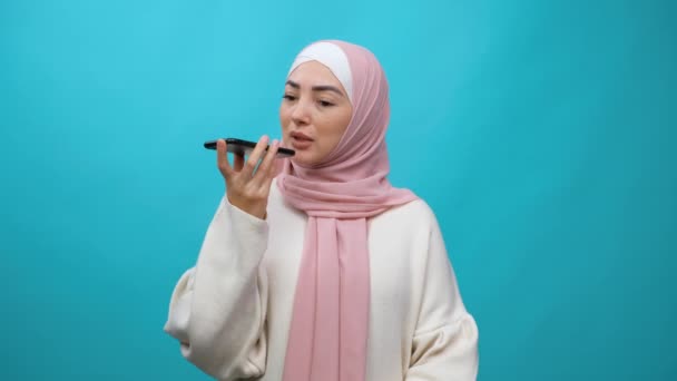 Muslim woman in hijab leaves a voice message on the mobile phone. Concept of technology, assistant help, communicate online. Isolated studio shot on blue background — Stock Video