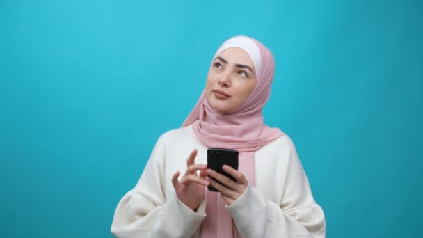 Glücklich schöne Studentin Junge muslimische Frau im Hidschab macht Notizen im Handy, denkt kreative Idee, Zukunftspläne und To-do-Liste, sieht inspiriert aus. Innenstudio isoliert auf blauem Hintergrund aufgenommen — Stockvideo