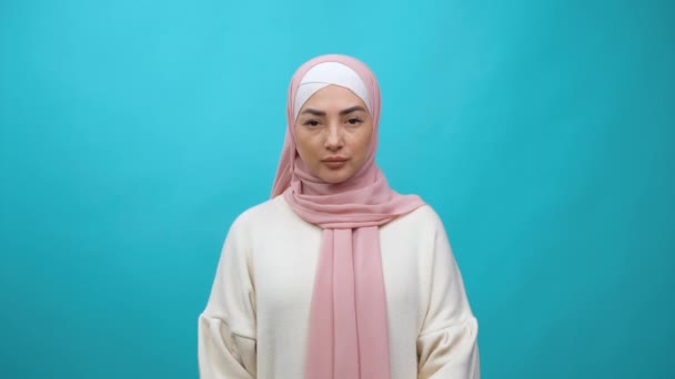 Potret wanita Muslim muda yang serius dalam jilbab berdiri tenang tanpa emosi, menatap kamera dengan penuh perhatian ekspresi tanpa senyum, benar-benar wajah termenung. pengambilan gambar studio dalam ruangan terisolasi pada latar belakang biru — Stok Video