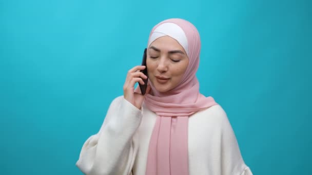 Sonriente joven musulmana en hijab usando hablar hablando por teléfono celular llevando a cabo una agradable conversación Estudio aislado filmado sobre fondo azul. — Vídeo de stock
