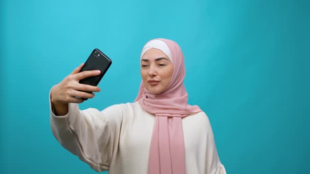 年轻的穆斯林妇女在头巾采取自拍。蓝色背景的孤立演播室拍摄 — 图库视频影像