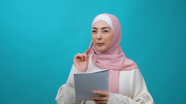Счастливая красивая молодая мусульманка в хиджабе делает заметки в бумажном блокноте, пишет творческую идею, планы на будущее и список дел, выглядит вдохновленной. Изолированная студия — стоковое видео
