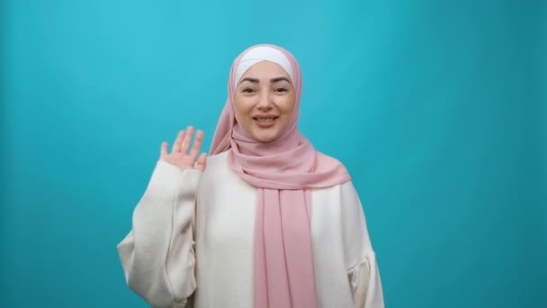 Perempuan Muslim muda yang ramah dalam hijab tersenyum ramah di depan kamera dan melambaikan tangan isyarat halo atau selamat tinggal, menyambut dengan ramah ekspresi. Rekaman studio yang terisolasi — Stok Video