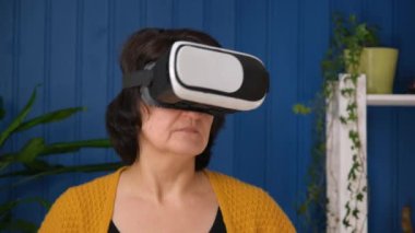 Orta yaşlı kadın oturma odasında sanal gerçeklik gözlüğü kullanarak modern oyunlarda oynuyor. Çevrimiçi eğitim ve evdeki metaevren geleceğin VR AR modern teknolojilerini kullanıyor..