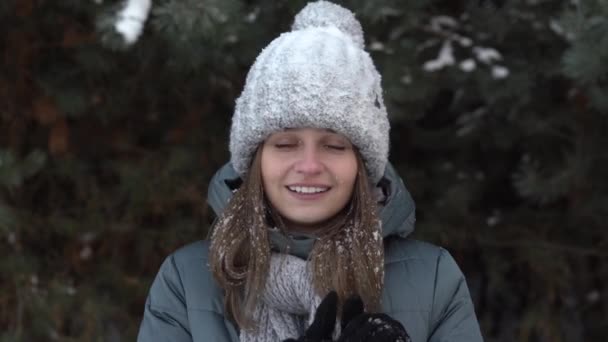 Ένα όμορφο κορίτσι με χειμωνιάτικα ρούχα σκουπίζει το πρόσωπό της από το χιόνι στο φόντο του δάσους. — Αρχείο Βίντεο