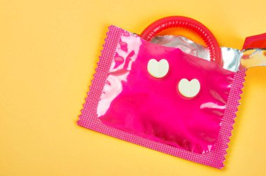 Sarı arka planda kalp şeklindeki ilaçla açılmış pembe prezervatif paketi. Güvenli seks ve üreme sağlığı kavramı.