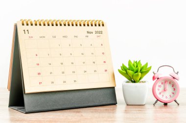 November 2022 Desk calendar with pink alarm clock on wooden table.