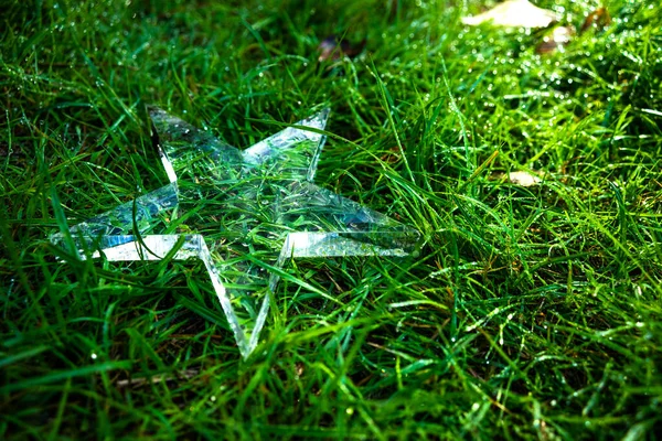 A transparent ice star lies on the ground in the grass. A glass cup in the form of a star. — Stockfoto