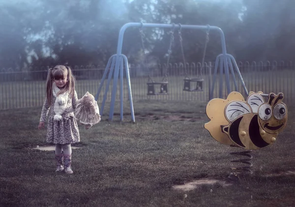 Жуткая, туманная игровая площадка с маленькой девочкой, держащей плюшевого мишку — стоковое фото