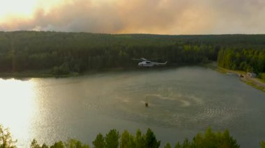 İtfaiyeci helikopteri, orman yangınını tasfiye etmek için göletteki kepçeyle su çekiyor