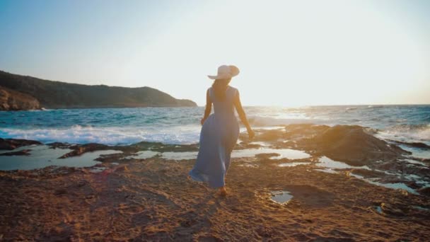 Junges Mädchen im Sommerkleid läuft barfuß am felsigen Strand, der von Meereswellen angespült wird — Stockvideo