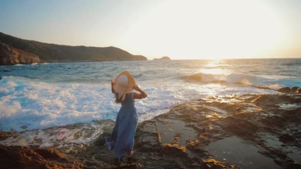 Młoda kobieta w letniej sukience stojąca na skalistej plaży umyta falą morską — Wideo stockowe