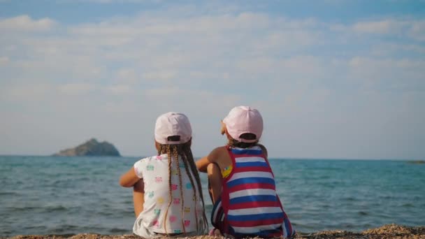 Två små flickor sitter på stranden, tittar till havs, barn på semester resor — Stockvideo