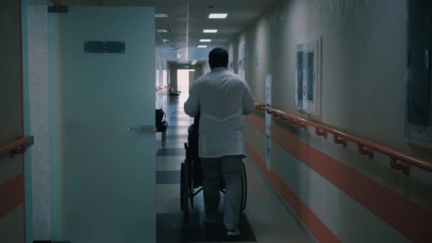 Krankenschwester schiebt Rollstuhl mit behindertem Patienten durch dunklen Krankenhausflur — Stockvideo