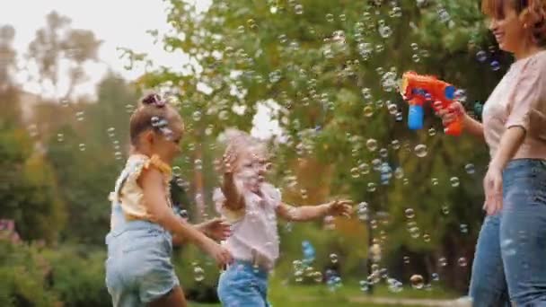 Щаслива мати дме мильні бульбашки з іграшковим пістолетом, маленькі дочки намагаються зловити — стокове відео