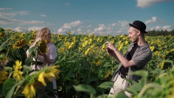 Blond flicka i ett solrosfält läser bok, poserar på telefonen till söt kille — Stockvideo